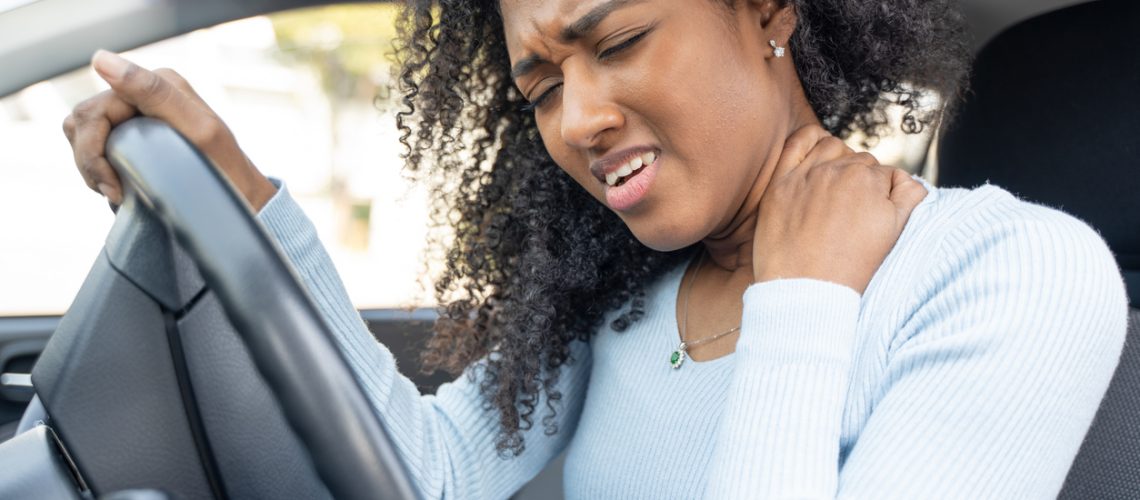 A black woman inside a car rubbing her neck in pain.